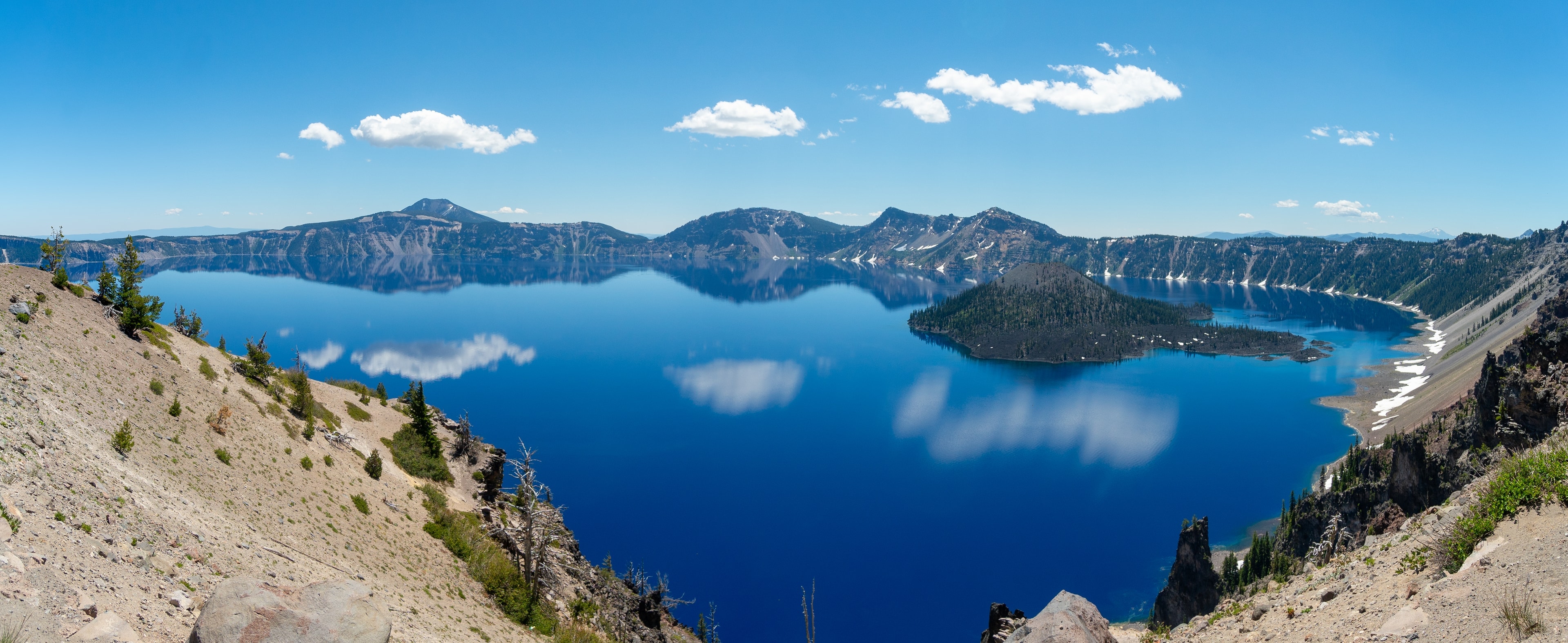 A wide view of Crater Lake and Wizard Island from the rim in July, white clouds over the far wall