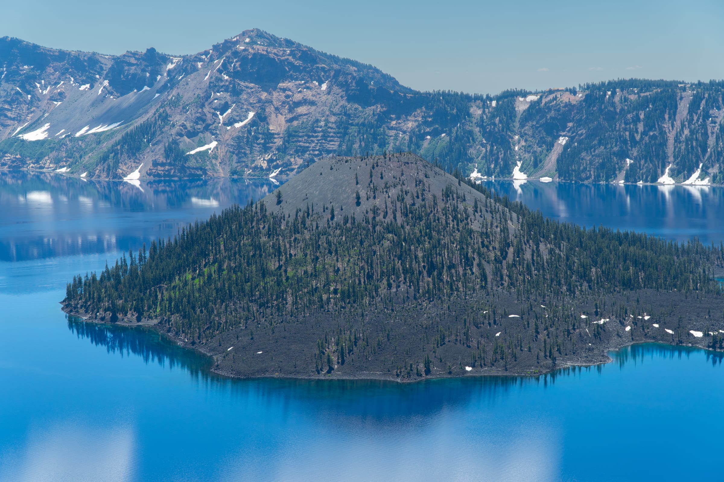 Wizard Island close up in the blue water of Crater Lake in July, snow still on the caldera walls behind